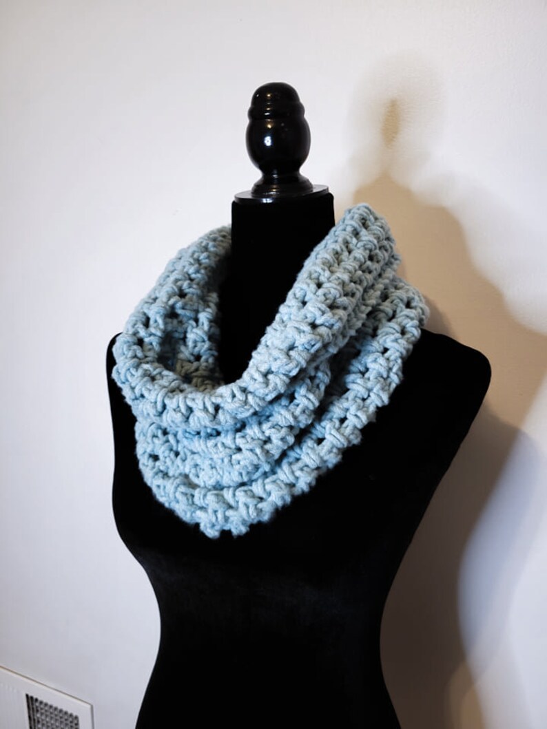 A light blue crochet fleece cowl displayed on a black velvet mannequin against a white backdrop at a side angle.