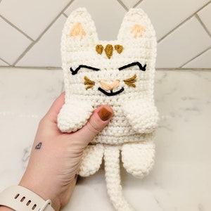 A hand holding the cream and caramel crochet kitty cat for size reference against a tile background. The kitten has half moon eyes and a smile with whiskers.