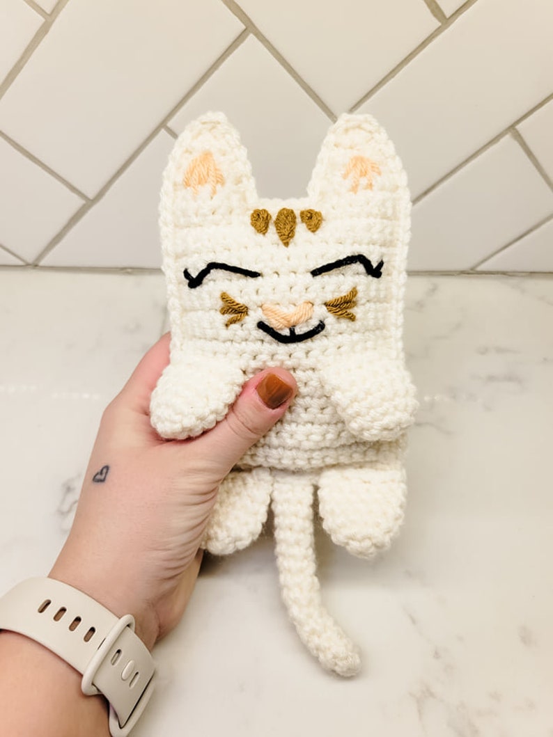 A hand holding the cream and caramel crochet kitty cat for size reference against a tile background. The kitten has half moon eyes and a smile with whiskers.