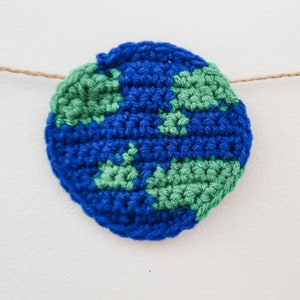 A crocheted planet earth is strung across a white background with jute. You can see each stitch made with blue and green yarn.