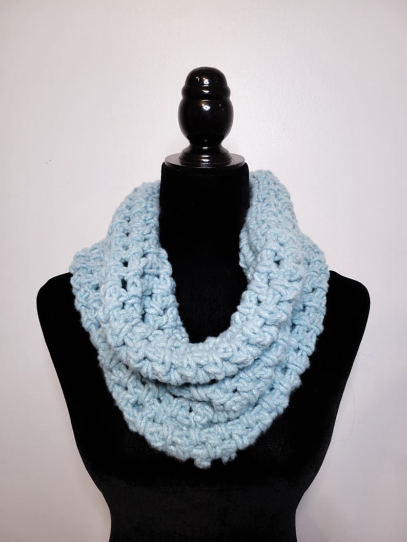 A light blue crochet fleece scarf displayed on a black velvet mannequin against a white backdrop.