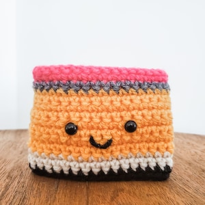 A crocheted coffee cozy in the layered colors of a pencil sits on a wooden table top with a white background. The cozy has wideset eyes and a tiny smiling mouth.