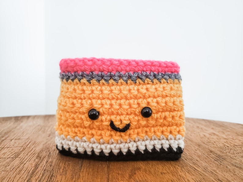 A crocheted coffee cozy in the layered colors of a pencil sits on a wooden table top with a white background. The cozy has wideset eyes and a tiny smiling mouth.