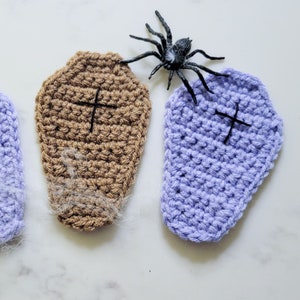 Two crochet coffin coasters, one purple and one brown with spider webs and a spider on a marble countertop.