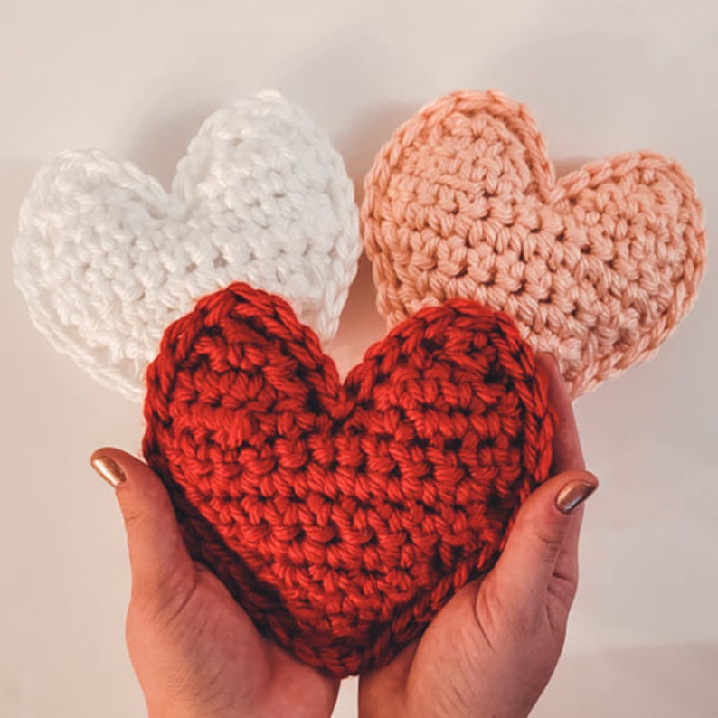 A set of three large crochet heart plushies made of bulky / chunky yarn against a white backdrop. There is one pink, one red being held by hands for size reference, and one white heart.