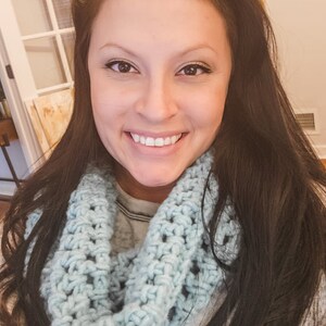 A smiling girl with long, brunette hair wearing the light blue crochet fleece cowl / scarf.