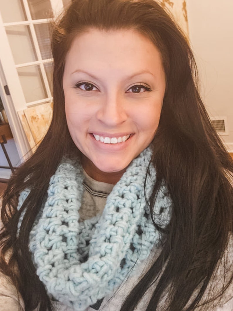 A smiling girl with long, brunette hair wearing the light blue crochet fleece cowl / scarf.
