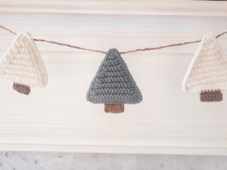 Crocheted Christmas Trees strung into a garland draped across a white mantle with jute. The trees are made of green and cream yarn with light brown trunks.