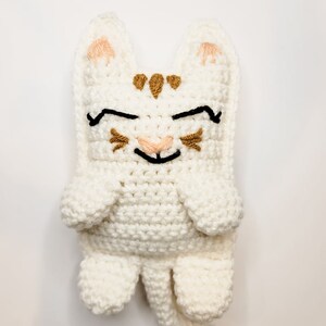 The cream and caramel crochet kitty cat against a white background laying flat. The kitten has half moon eyes and a smile with whiskers, a long tail and pointed ears.