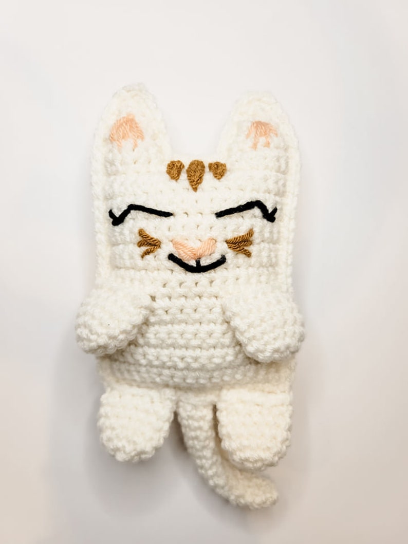 The cream and caramel crochet kitty cat against a white background laying flat. The kitten has half moon eyes and a smile with whiskers, a long tail and pointed ears.