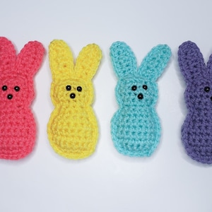 Four small crocheted peeps sit in a row along a white background. From left to right their colors are pink, yellow, blue and purple. Each has two tiny dots for eyes and one tiny dot nose.