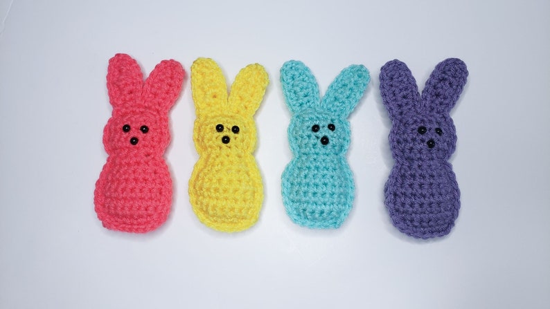 Four small crocheted peeps sit in a row along a white background. From left to right their colors are pink, yellow, blue and purple. Each has two tiny dots for eyes and one tiny dot nose.