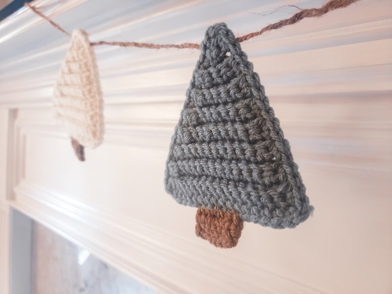 Crocheted Christmas Trees strung into a garland draped across a white mantle with jute. The trees are made of green and cream yarn with light brown trunks.