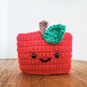 A crocheted coffee cozy in the colors of an apple sits on a wooden table top with a white background. The cozy has wideset eyes and a tiny smiling mouth. A tiny stem and leaf are at the top.