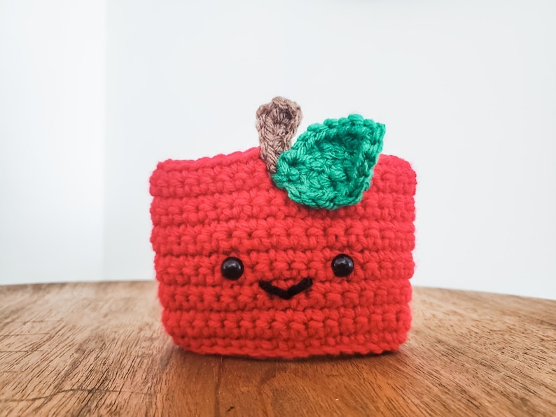 A crocheted coffee cozy in the colors of an apple sits on a wooden table top with a white background. The cozy has wideset eyes and a tiny smiling mouth. A tiny stem and leaf are at the top.