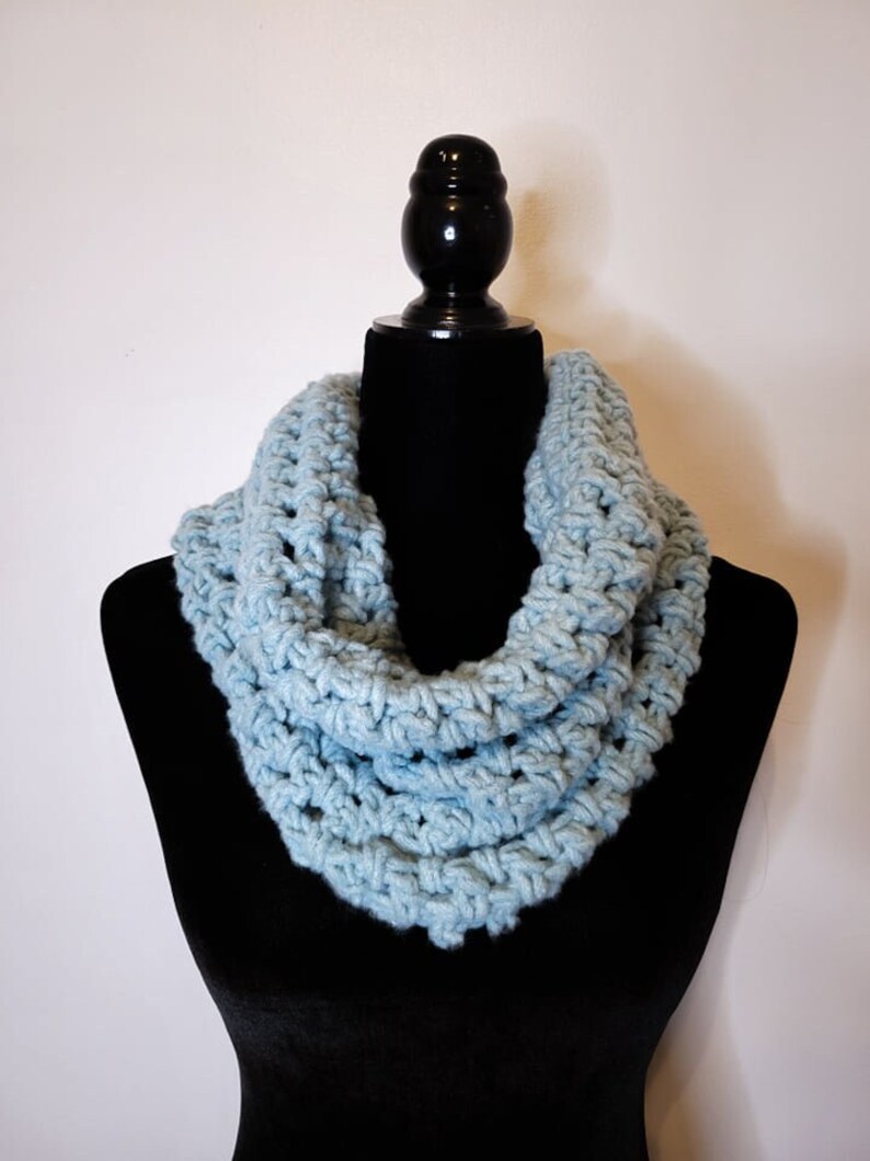 A light blue crochet fleece cowl displayed on a black velvet mannequin against a white backdrop.