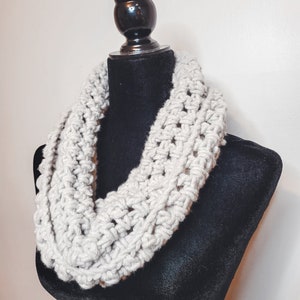 A light grey crochet fleece cowl displayed on a black velvet mannequin against a white backdrop.