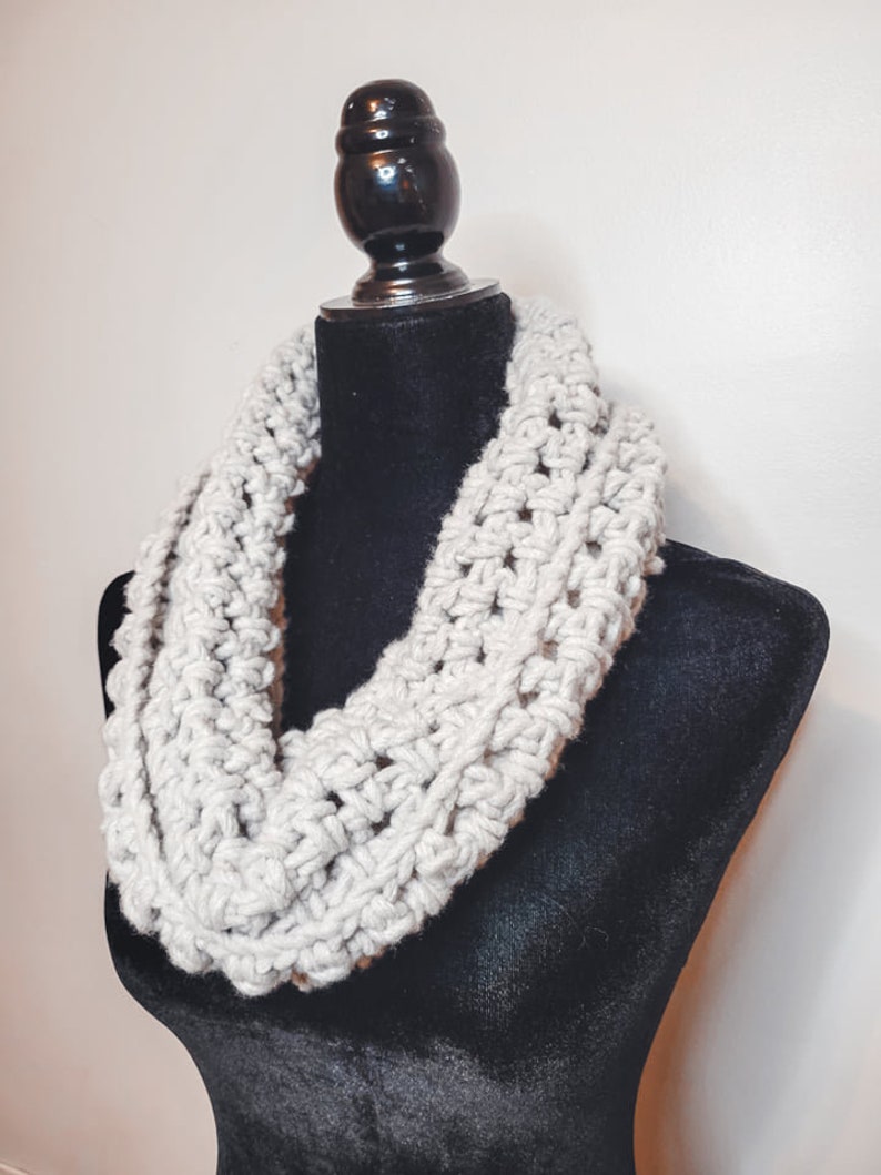 A light grey crochet fleece cowl displayed on a black velvet mannequin against a white backdrop.