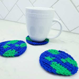 A crocheted planet earth is displayed as a coaster with a white ceramic mug atop. You can see each stitch made with blue and green yarn on the planet coaster next to it on a white marble counter top.