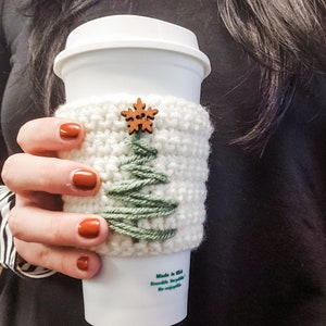 The crochet Christmas Tree Cozy in use on a white Starbucks travel mug being held by a hand with painted nails.