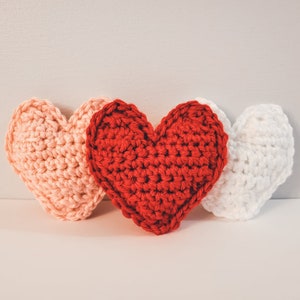 A set of three large crochet hearts made of bulky / chunky yarn against a white backdrop. There is one pink, one red, and one white heart.