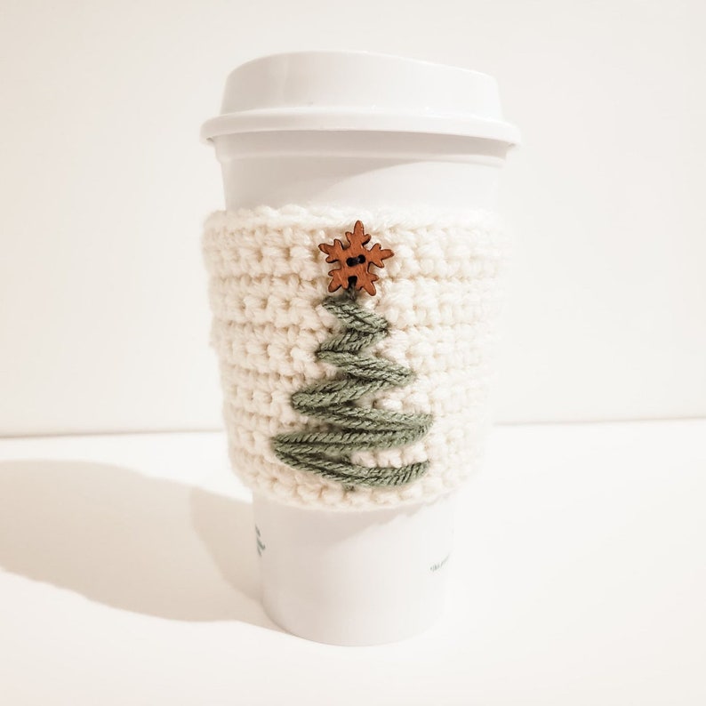 A crocheted Christmas Tree coffee cozy displayed on a white Starbucks mug with a white background. The cup cozy is minimalist using green, cream and wood.
