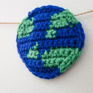 A crocheted planet earth is strung across a white background with jute. You can see each stitch made with blue and green yarn.