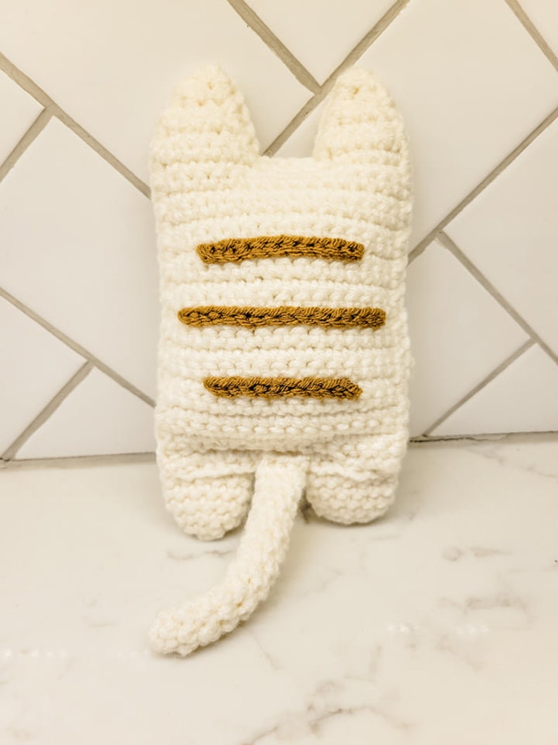 The backside view of the crochet kitty cat against a white background displaying three caramel colored stripes on the kitty's back.