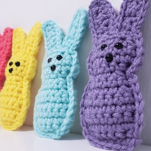 Four small crocheted peeps sit in a row along a white background. From left to right their colors are pink, yellow, blue and purple. The purple is closest to the viewer in this side angle image.