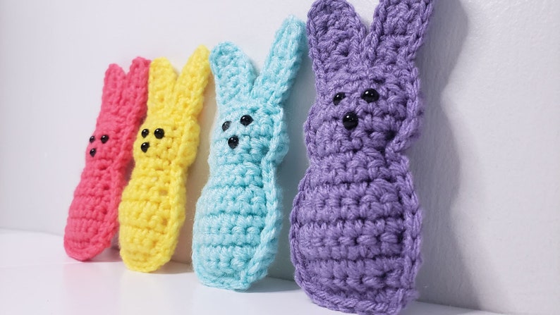 Four small crocheted peeps sit in a row along a white background. From left to right their colors are pink, yellow, blue and purple. The purple is closest to the viewer in this side angle image.