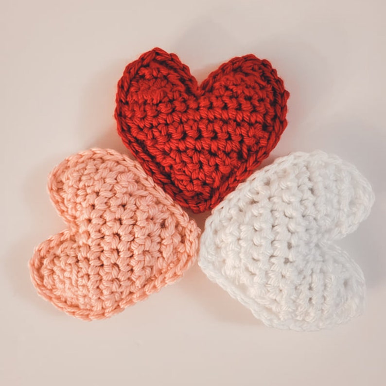 A set of three large crochet heart plushies made of bulky / chunky yarn against a white backdrop displayed in a semicircle. There is one pink, one red, and one white heart.