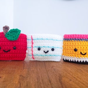 On a wooden table top from left to right is a piece of paper crochet cozy, a pencil and an apple. Each cozy is hand crocheted with yarn and has a smiling little face.