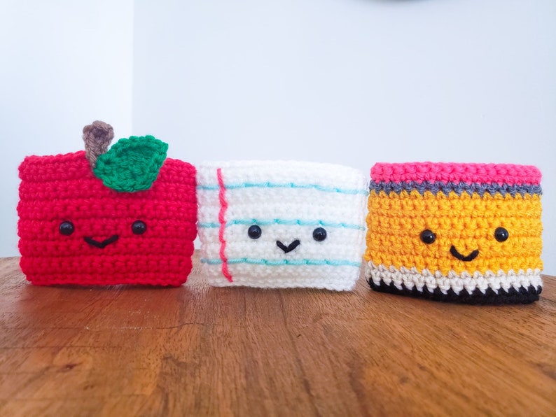 On a wooden table top from left to right is a piece of paper crochet cozy, a pencil and an apple. Each cozy is hand crocheted with yarn and has a smiling little face.