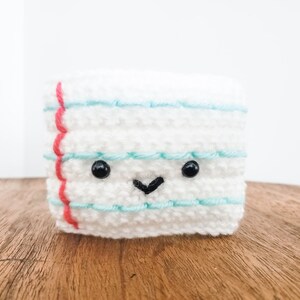 A crocheted coffee cozy in the layered colors of a piece of ruled paper sits on a wooden table top with a white background. The cozy has wideset eyes and a tiny smiling mouth.
