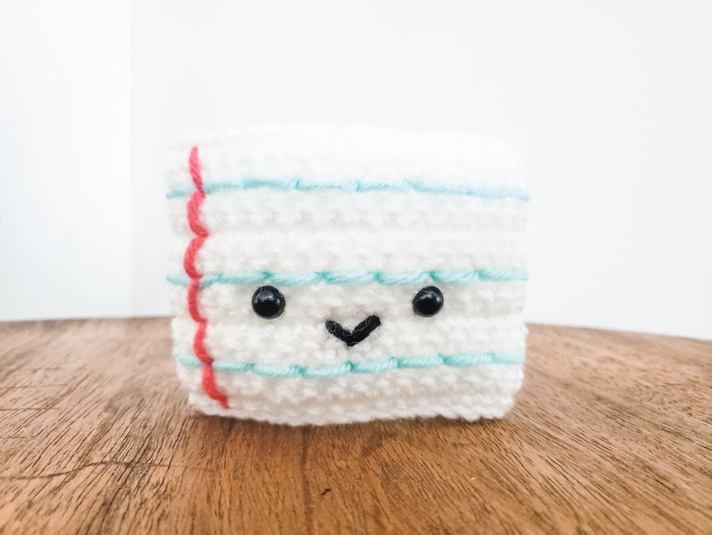 A crocheted coffee cozy in the layered colors of a piece of ruled paper sits on a wooden table top with a white background. The cozy has wideset eyes and a tiny smiling mouth.