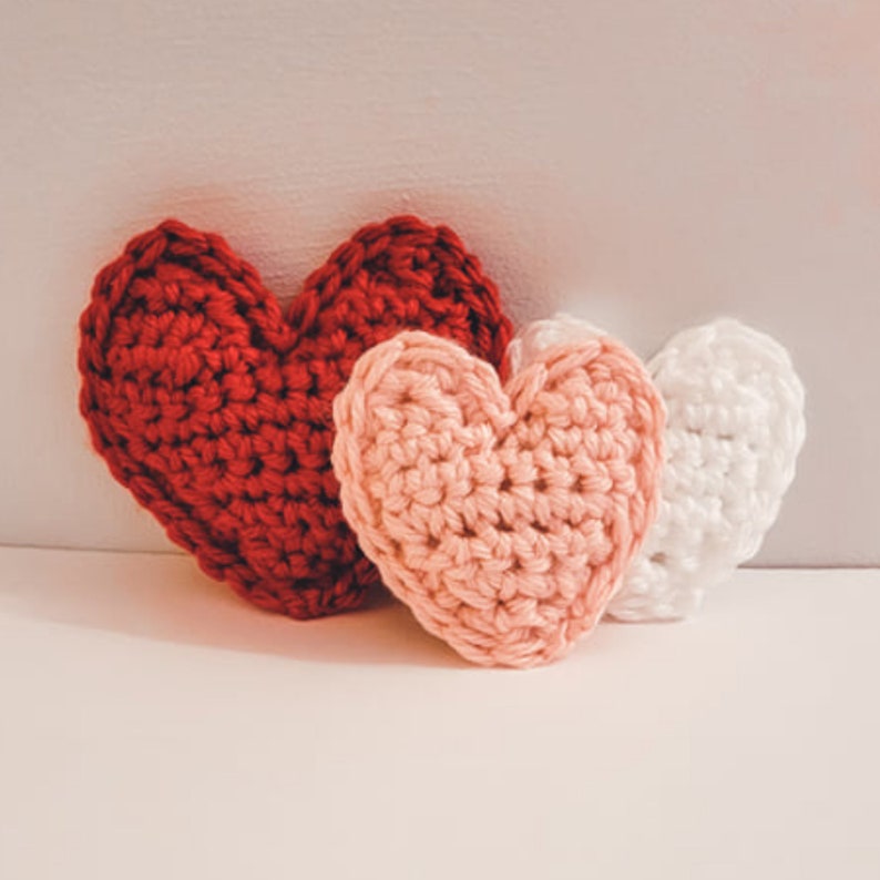 A set of three crochet heart plushies made of bulky / chunky yarn against a white backdrop. There is one small pink, one large red, and one small white heart pillow.