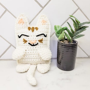 The cream and caramel crochet kitty cat against a tile background with a small green plant. The kitten has half moon eyes and a smile with whiskers, a long tail and pointed ears.