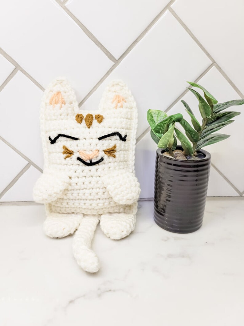 The cream and caramel crochet kitty cat against a tile background with a small green plant. The kitten has half moon eyes and a smile with whiskers, a long tail and pointed ears.