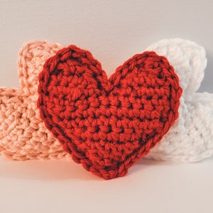 A red crochet heart pillow standing upright with a pink and white laying to either side against a white background.