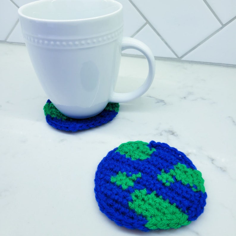 A crocheted planet earth is displayed as a coaster with a white ceramic mug atop. You can see each stitch made with blue and green yarn on the planet coaster next to it on a white marble counter top.