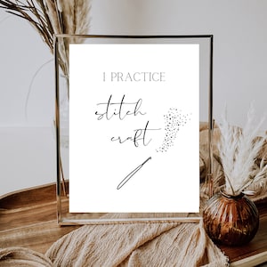 May include: A framed print with the text "I practice stitch craft" and a stylized needle and thread design. The print is in a gold frame and is sitting on a wooden surface.