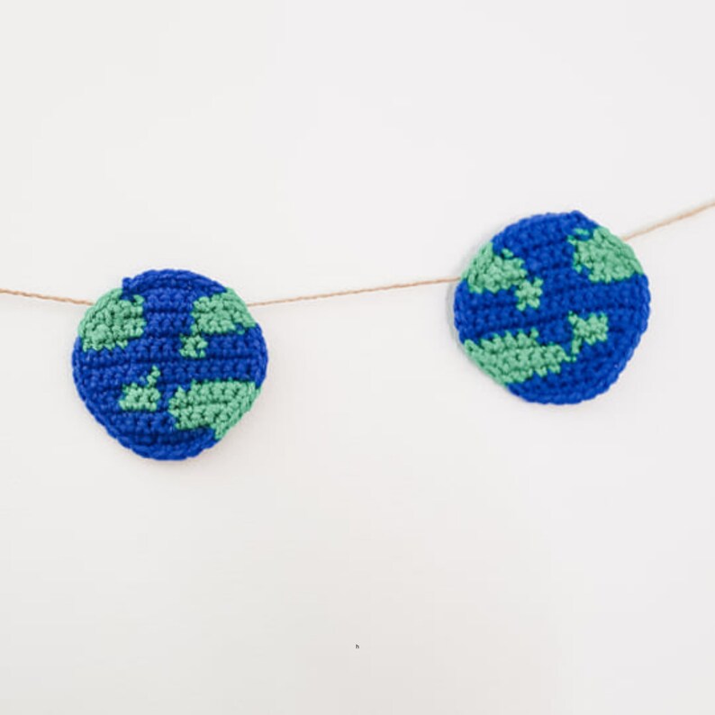 Two crocheted planet earths are strung across a white background with jute. You can see each stitch made with blue and green yarn.