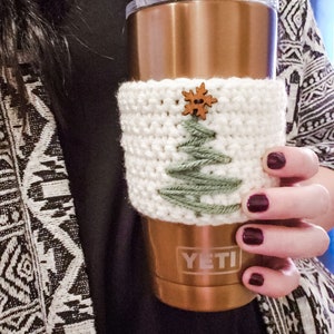 An up close image of the crocheted Christmas Tree coffee cozy displayed in hand on a gold yeti mug. The cup cozy is minimalist using green and cream yarn and a wooden button.