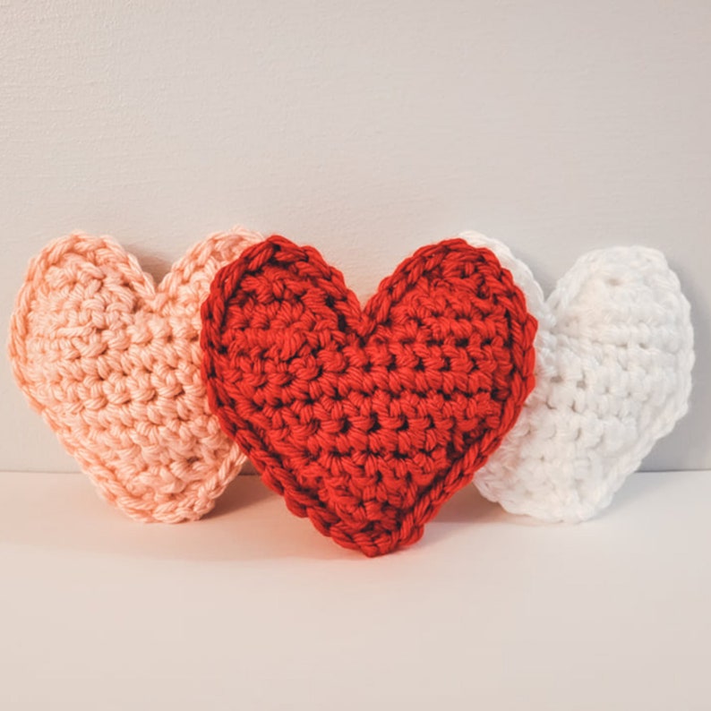 A set of three large crochet heart plushies made of bulky / chunky yarn against a white backdrop. There is one pink, one red, and one white heart.