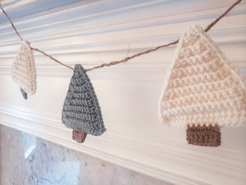 Crocheted Christmas Trees strung into a garland draped across a white mantle with jute. The trees are made of green and cream yarn with light brown trunks.