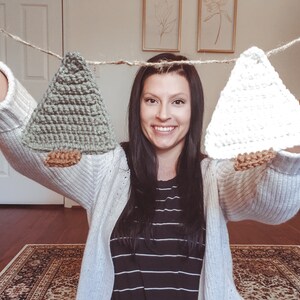 Crocheted Christmas Trees strung into a garland being held in hand by the artist. The trees are made of green and cream yarn with light brown trunks string together with jute.