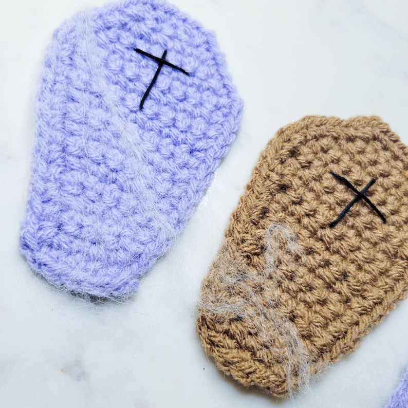 Two crochet coffin coasters, one purple and one brown with spider webs and a spider on a marble countertop.