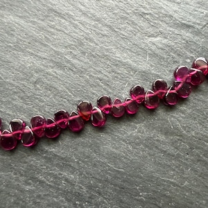 May include: A string of deep magenta-coloured, teardrop-shaped beads. The beads are strung together, creating a necklace or bracelet. The beads have a translucent quality, allowing light to pass through.