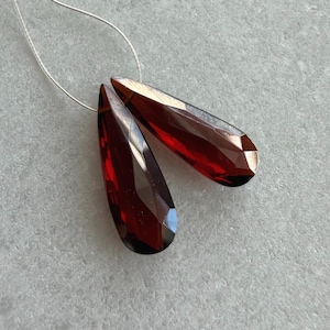 May include: Two faceted, teardrop-shaped, deep red gemstones are strung on a thin, silver-colored wire. The gems have a polished, reflective surface, catching the light and displaying a range of dark red hues. The background is a light gray, textured surface.
