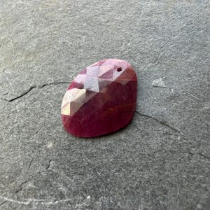 May include: A faceted, oval-shaped ruby gemstone with a deep red hue. The gemstone has a small hole drilled at the top, suggesting it could be used as a pendant or bead. The surface reflects light, showcasing its cut.